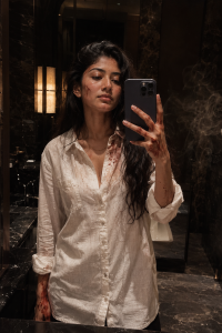Ultra realistic cinematic mirror selfie of a powerful young Indian man standing in a dark luxury hotel bathroom at night, messy textured hair, sharp jawline, cold confident expression, oversized white linen shirt partially unbuttoned with sleeves rolled up, subtle gold chain and luxury watch, elegant mafia boss aura, faint realistic blood stains and minor scratches on collar, knuckles and face, dramatic warm ambient lighting, glossy marble interiors, deep cinematic shadows, soft smoke haze near the mirror, intense late-night tension, realistic skin texture, emotionally detached expression, luxury Tamil action-movie aesthetic, raw masculine energy, classy but dangerous vibe, subtle film grain, high-detail editorial photography, stylish action-scene aftermath vibe, realistic reflection, 35mm cinematic composition